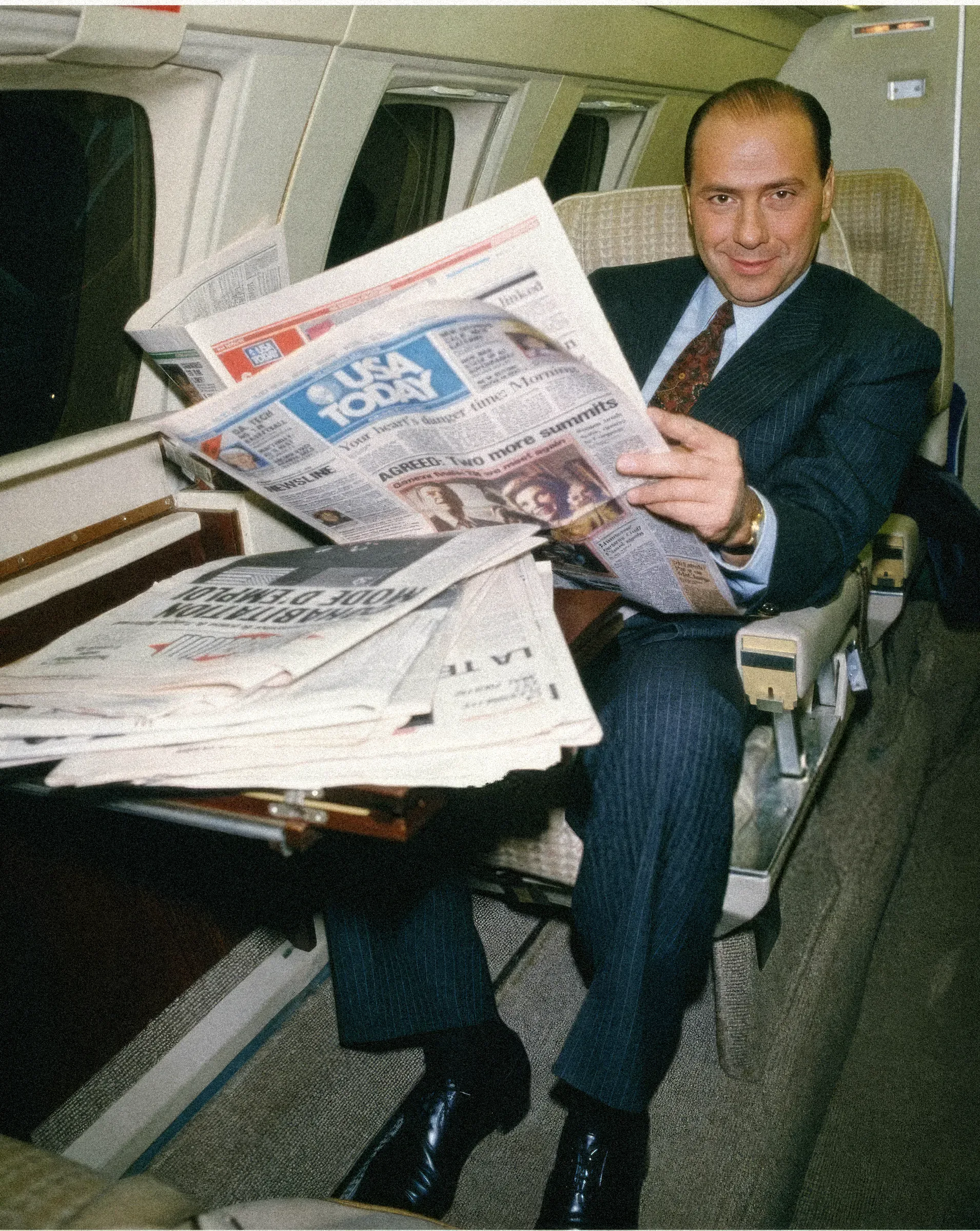 Un uomo in completo legge un giornale in aereo, con altri quotidiani sulle ginocchia.